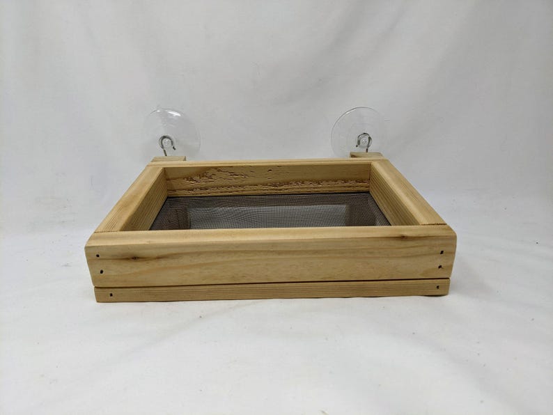 May include: A wooden bird feeder with a mesh bottom and two suction cups for attaching to a window. The feeder is made of natural wood and has a simple design.