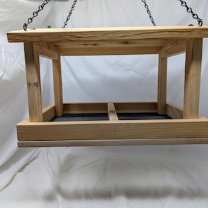 Puede incluir: Un comedero para p&aacute;jaros de madera con una cadena de metal negro colgando de la parte superior. El comedero tiene una forma rectangular con una parte superior plana y una parte inferior que est&aacute; dividida en dos secciones. El comedero est&aacute; hecho de madera natural de color marr&oacute;n claro.