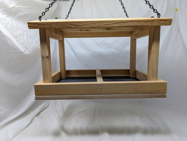 Puede incluir: Un comedero para p&aacute;jaros de madera con una cadena de metal negro colgando de la parte superior. El comedero tiene una forma rectangular con una parte superior plana y una parte inferior que est&aacute; dividida en dos secciones. El comedero est&aacute; hecho de madera natural de color marr&oacute;n claro.