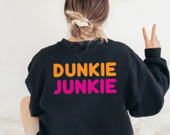 以下が含まれることがあります： オレンジとピンクの文字で「Dunkie Junkie」と書かれた黒いスウェットシャツです。