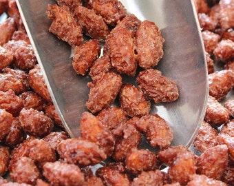 May include: A close-up of a metal scoop filled with sugared almonds. The almonds are a deep brown color and are coated in a layer of sugar crystals. The almonds are piled high in the scoop and are spilling out over the edges.