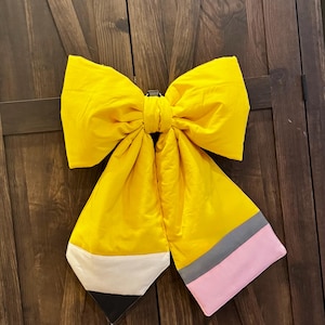 May include: A large, yellow bow shaped like a pencil, with a black, white, gray, and pink tip. The bow is made of a shiny fabric and is hanging against a dark brown wooden door.