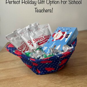 Puede incluir: Una cesta de tela azul y roja con un patrón de lunares y manzanas rojas impresas en ella. La cesta está llena de besos de chocolate, paquetes de chocolate caliente Swiss Miss y una pequeña caja azul con un diseño de Papá Noel y el texto "Russell Stover" en ella.