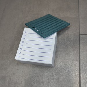 Op de afbeelding: Een wit notitieboekje met blauwe lijnen en een tealblauwe plastic checklist erop. De checklist heeft 10 vierkantjes.