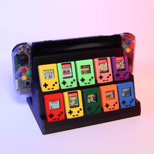 May include: A black display stand holding ten colorful miniature game cartridges. The cartridges are in various colors, including yellow, green, pink, and blue. A Nintendo Switch console is visible in the background.