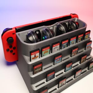May include: A grey plastic storage stand for a Nintendo Switch console, controllers, and game cartridges. The stand has multiple tiers for storing game cartridges and a top tier for the console and controllers. The stand is made of a textured plastic and has a grey colour.