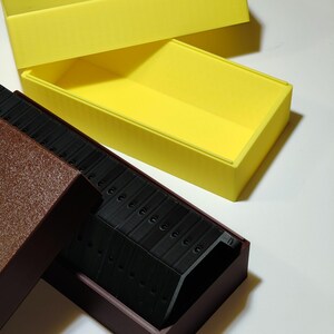 May include: A yellow rectangular box with a matching lid is open, revealing a dark brown box with a dark interior. Inside the brown box are several black, curved objects. The boxes are on a white surface.