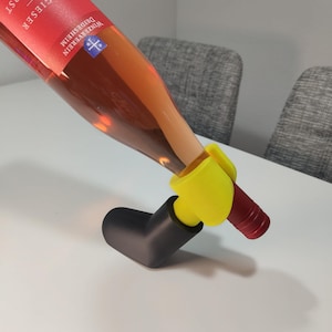 May include: A bottle of rosé wine is held by a yellow and black wine bottle holder. The wine bottle has a red label with white text. The holder is on a white surface.