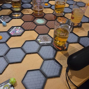 May include: A tabletop board game featuring interlocking hexagonal tiles with stone and wood patterns. Several clear glasses filled with golden liquid are placed on the tiles. Game components include a green twenty-sided die, a blue die, and character cards with illustrations and text like Absinthia of Wormwood and Poltergeist.