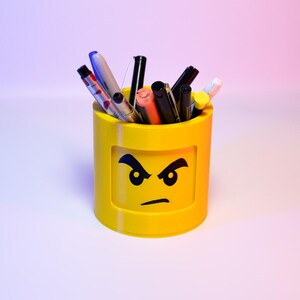 Puede incluir: Un portalápices cilíndrico amarillo con un diseño de cara enfadada. El portalápices está lleno de bolígrafos y rotuladores de varios colores. El diseño recuerda a un popular juguete de construcción.