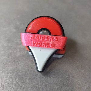May include: A red and white pin with a black border, featuring a pink banner with the text "RAIDERS WORLD." The pin has a circular top and a white, triangular bottom, resembling a location marker.