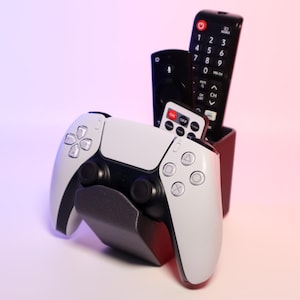 Könnte beinhalten: Ein weiß-schwarzer Videospiel-Controller mit einem schwarzen und silbernen Ständer. Der Ständer hält drei schwarze Fernbedienungen. Der Controller hat Knöpfe mit den Beschriftungen "Dreieck", "Quadrat", "Kreis" und "Kreuz".