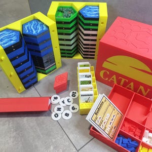 Op de afbeelding: Een 3D-geprinte organizer voor het bordspel Catan. De organizer bevat vakken voor de tegels, kaarten en andere componenten van het spel. De organizer is geel en rood met het woord "CATAN" op de voorkant.