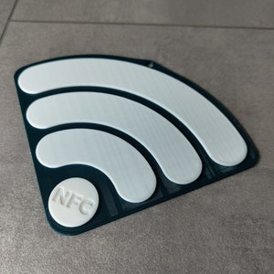 Peut inclure: Une étiquette NFC en plastique vert foncé avec un symbole wifi blanc et un cercle blanc avec le texte "NFC" au centre.