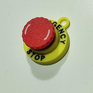 Può includere: Un pulsante di arresto di emergenza rosso e giallo. Il pulsante ha una parte superiore rossa strutturata e una base gialla con le parole "STOP" e "EMERGENCY" stampate in nero. Ha un piccolo anello laterale.