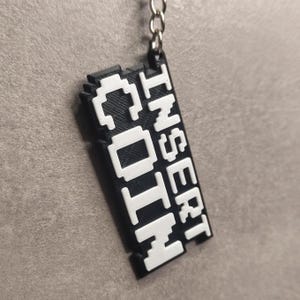 May include: Black and white pixelated keychain with the text "Insert Coin" and "Goth" in white.
