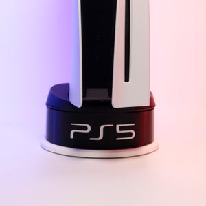 Könnte beinhalten: Eine weiße und schwarze PlayStation 5 Konsole mit einem schwarzen Sockel, auf dem "PS5" in weißen Buchstaben steht.