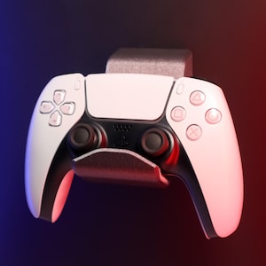 Op de afbeelding: Een witte PlayStation 5-controller met zwarte knoppen en analoge sticks is gemonteerd op een zwarte wandbeugel. De controller is naar voren gericht en de knoppen zijn zichtbaar.