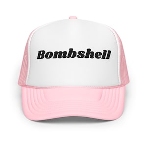May include: A pink and white trucker hat with the word "Bombshell" in black, bold lettering across the front. The hat features a pink brim and mesh backing, with a white front panel. The hat has a curved brim and a pink button on top.