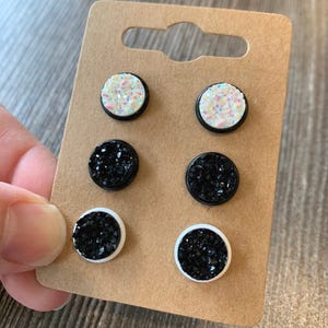 May include: A set of six stud earrings with black and white glitter. The earrings are displayed on a brown card.