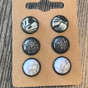 May include: Six pairs of black and white stud earrings on a brown card. The earrings have different patterns, including marble, speckled, and cracked.