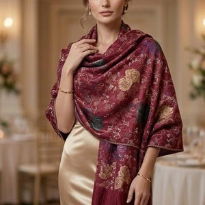 May include: A deep burgundy shawl featuring a woven floral pattern in gold and dark green tones, finished with delicate fringe along the bottom edge. The shawl is draped over a smooth, sleeveless champagne-colored satin dress.
