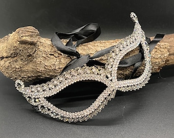 Puede incluir: Una máscara plateada para mascaradas adornada con brillantes pedrería. La máscara presenta un diseño intrincado con una forma curva y está acentuada con cintas de satén negro para atar. La máscara se exhibe sobre un trozo de madera.
