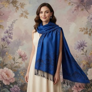 May include: A woman wearing a royal blue shawl with a black paisley design and fringe. The shawl is draped over a cream-colored dress. The background features a floral mural with pink, purple, and white flowers.