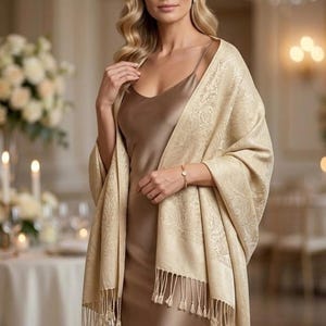 May include: A champagne-colored pashmina shawl featuring an intricate tonal paisley pattern and long fringe trim, draped over a smooth, sleeveless brown satin slip dress with a side slit and a thin spaghetti strap neckline.