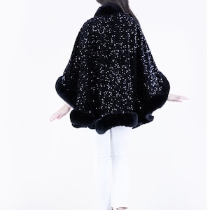 May include: A black sequined cape with a fur trim. The cape is worn by a person with long dark hair and white pants.