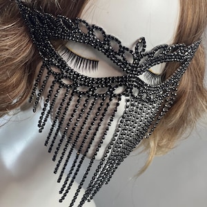 May include: A black masquerade mask with a delicate, intricate design. The mask is adorned with rhinestones and features a long fringe of dangling beads.