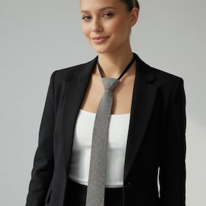 May include: A silver-toned, crystal-studded tie with a black neckband. The tie is worn with a black blazer and a white top. The image is a medium shot, with a neutral background.