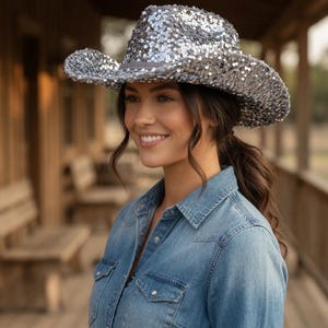 May include: A silver sequin cowboy hat with a gray brim. The hat is worn by a person wearing a denim shirt. The hat has a wide brim and a classic cowboy hat shape. The sequins give the hat a sparkly appearance.