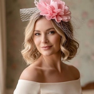 May include: A pale pink floral headpiece with white feathers and a white net veil. The headpiece is worn by a person wearing an off-the-shoulder cream dress. The background features a soft, blurred floral pattern.
