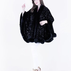 May include: A black sequined cape with a fur trim. The cape is worn by a model with white pants and heels.
