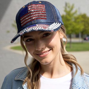 May include: Denim baseball cap featuring an American flag design. The flag is embellished with red and silver rhinestones, with silver rhinestones along the brim. The cap is a dark blue color.