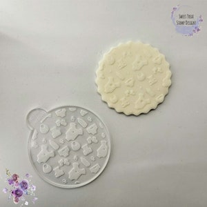 May include: A clear, circular stencil featuring baby-themed designs, such as onesies, bottles, and dummies, next to a white, scalloped-edge biscuit with matching embossed patterns. The text "Sweet Treat Stamp Designs" is visible.