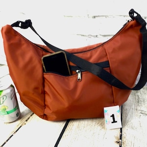 May include: A rust-colored nylon crossbody bag with a black adjustable strap. The bag has a zippered pocket on the front and a smaller pocket on the side that can hold a phone. The bag is sitting on a white wooden surface.