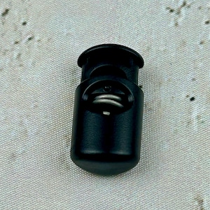 May include: A black plastic toggle with a spring mechanism. The toggle is designed to be used with cords or straps.