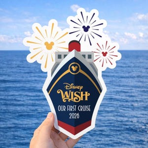 May include: A Disney Wish cruise ship-shaped sticker with the text "Disney Wish OUR FIRST CRUISE 2026." The sticker is held in front of a blue ocean background. The sticker has a navy blue and gold design with red accents and Mickey Mouse fireworks.