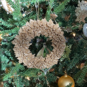 May include: A gold glitter Christmas wreath ornament hanging on a green Christmas tree.