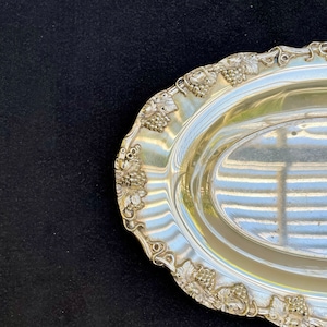 May include: A silver oval serving dish with a decorative grapevine design around the rim.