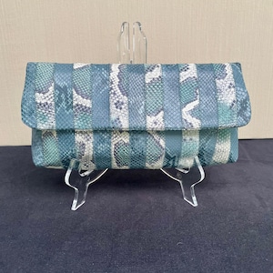 May include: A green and white striped clutch purse with a snakeskin pattern. The purse is on a clear acrylic stand.