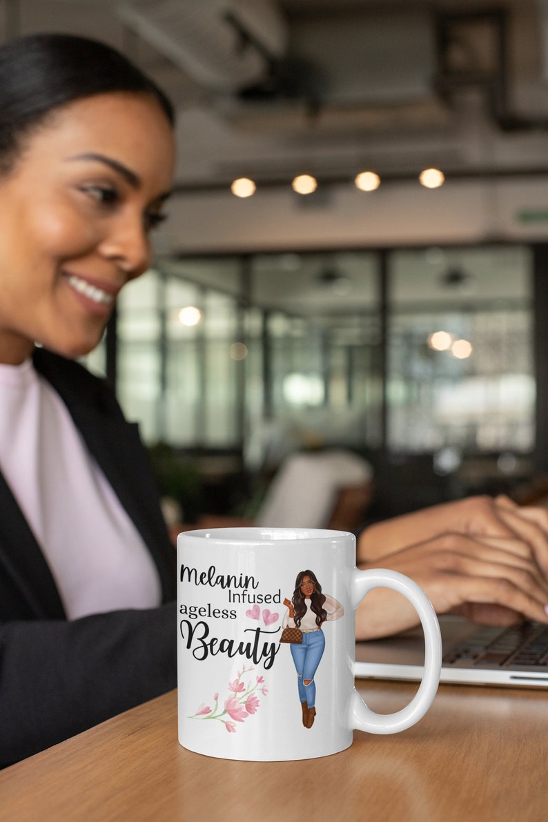 May include: White ceramic mug with a black and white illustration of a woman with long black hair, wearing a white shirt, blue jeans, and brown boots. The mug has the text "Melanin infused ageless Beauty" with pink hearts and pink flowers.