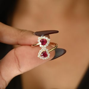 Może przedstawiać: Złoty pierścionek z dwoma czerwonymi kamieniami w kształcie serca, każdy otoczony aureolą małych, przezroczystych kamieni. Pierścionek ma delikatny złoty pierścień i elegancki design.