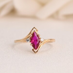 May include: A gold ring with a marquise-cut ruby gemstone, and two smaller white gemstones. The ring has a delicate, swirling design with a V-shaped setting around the ruby. The ring is set against a soft, white background.