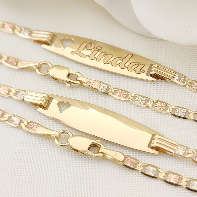 K&ouml;nnte beinhalten: Drei goldene Armb&auml;nder mit einer zarten, diamantgeschliffenen Kette. Das mittlere Armband hat ein personalisiertes Namensschild mit dem Namen "Linda", der in Kursivschrift graviert ist. Jedes Namensschild hat einen kleinen Herz-Ausschnitt.