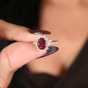 Può includere: Un anello d'oro con un grande rubino ovale, circondato da un alone di diamanti rotondi più piccoli. La fascia dell'anello è anch'essa ornata di piccoli diamanti, creando un effetto scintillante. L'anello è presentato su uno sfondo sfocato.