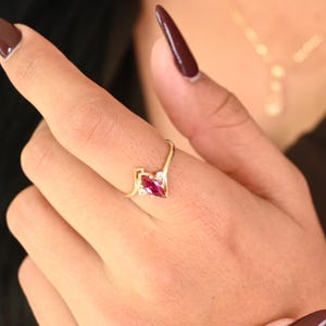 May include: A gold ring featuring a marquise-cut, deep red gemstone, flanked by two smaller white gemstones. The ring has a delicate, modern design, perfect for everyday wear or special occasions.