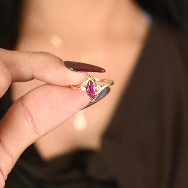 May include: A gold ring featuring a marquise-cut ruby gemstone, flanked by two smaller white gemstones. The ring has a delicate, openwork design and is held up against a blurred background.